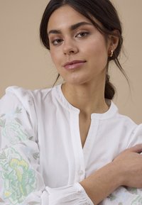 Jeune femme aux cheveux foncés portant un chemisier blanc avec des manches brodées de motifs floraux, regardant doucement l’appareil photo sur un fond beige.