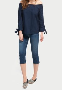 Marineblaue schulterfreies Oberteil mit langen Ärmeln und Schleifenakzenten an den Bündchen, kombiniert mit verkürzten blauen Jeans und gestreiften Ballerinas.