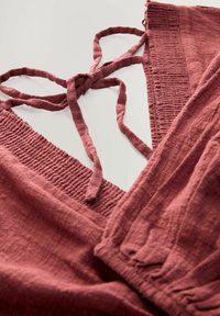Rust-colored linen garment with a textured, ruched bodice and tied straps, featuring a smooth fabric and loose-fit pants design.