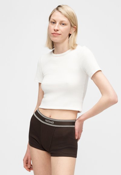 Ξανθιά γυναίκα με λευκό cropped T-shirt και μαύρο boxer Versace, στέκεται με το ένα χέρι στη μέση μπροστά σε απλό φόντο.