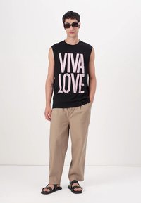 Svart ermeløs skjorte med "VIVA LOVE" i lys rosa, beige bukser og svarte sandaler. Glatt materiale, avslappet passform, minimalistisk design.