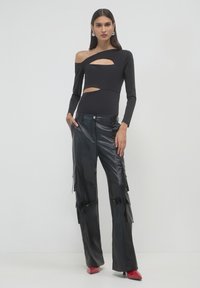 Donna che indossa un top nero a maniche lunghe con taglio asimmetrico, pantaloni cargo in finta pelle nera e tacchi rossi a punta, in piedi contro un muro bianco.