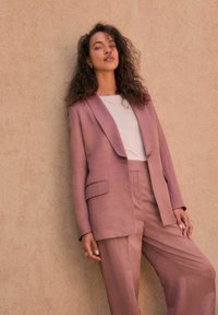 Dammig rosa linblazer med bred slag och ficka fram, matchande byxor. Slät textur och avslappnad passform.