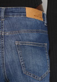 Μπλε τζιν παντελόνι με ίσια γραμμή, διαθέτοντας πίσω τσέπες και μια καφέ δερμάτινη ετικέτα στην ζώνη που αναγράφει "NOISY MAY".