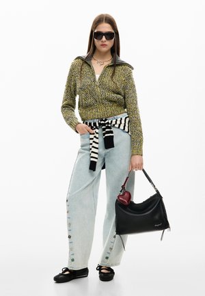 Naine, kes kannab päikeseprille, kollakasrohelist kootud lukuga kampsunit, helesiniseid nupuga laia säärega pükse, vöökohal seotud triibulist kampsunit, hoides musta käekotti.