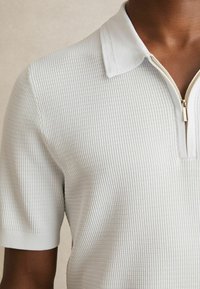 Nahaufnahme einer Person, die ein weißes, strukturiertes Kurzarm-Polohemd mit einem goldenen Reißverschluss trägt, der am Kragen teilweise geöffnet ist.