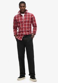 Camisa de cuadros roja y blanca con mangas largas, cuello abotonado, combinada con pantalones negros y zapatillas negras. Reloj visible en la muñeca.