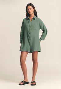 Zelený overal so zapínaním na gombíky vyrobený z ľahkej textílie, s dlhými rukávmi, vo voľnom strihu, s náprsníkovou kapsou a dekoratívnymi gombíkmi. Čierne sandále dopĺňajú vzhľad.