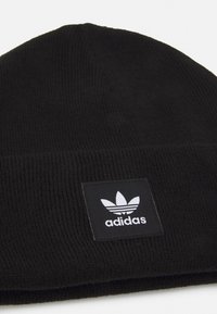 Zwarte gebreide beanie met een gestructureerd ribpatroon en een vierkante zwarte patch met een wit Adidas-logo. Eenvoudig en functioneel ontwerp.