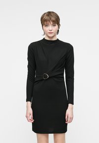 Mulher com cabelo castanho curto, vestindo um vestido preto justo de mangas longas com uma cintura franzida e uma fivela de metal circular, de pé contra um fundo branco.