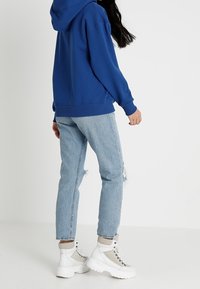Sudadera con capucha azul, jeans desgastados de un azul claro, zapatillas blancas gruesas con acentos grises. Vista trasera, mostrando un ajuste relajado y un estilo casual.