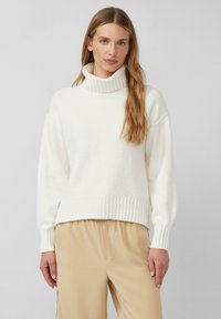Cremfarbener Rollkragenpullover mit gerippten Abschlüssen und einem weichen, strukturierten Stoff. Leicht oversized Fit, kombiniert mit beigen Hosen.