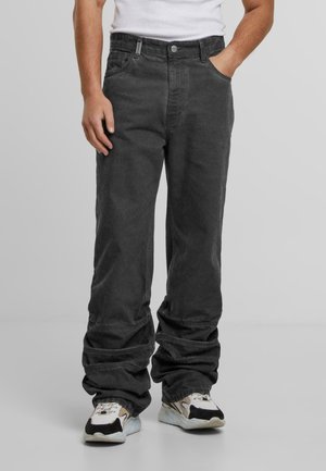 Homme portant un pantalon noir ample et des baskets blanches, se tenant avec une main dans la poche devant un fond uni.