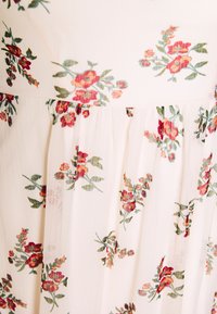 Blumenkleid aus leichtem Stoff, mit einem cremefarbenen Grundton und roten sowie grünen Blumenmustern sowie plissierten Details.