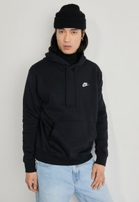 Mustaherra Nike-huppari, jossa on etutasku, säätönyöri hupussa ja valkoinen logo. Yhdistetty vaaleansinisiin farkkuihin ja mustaan neulottuun pipoon.