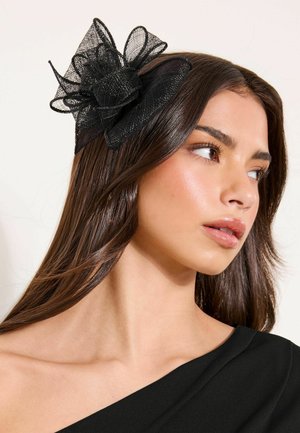 Noorukkene pruunide pikkade juustega naine, kes kannab musta õlgadeta pluusi ja suurt musta võrgust juuksenõela.