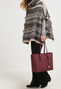 Vinrød taske i imiteret læder med to skulderstropper, gulddetaljer og præget logo; kombineret med en frynset, stribet poncho og sorte bukser.