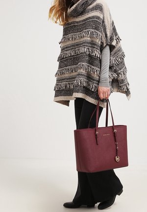 Vinrød taske i imiteret læder med to skulderstropper, gulddetaljer og præget logo; kombineret med en frynset, stribet poncho og sorte bukser.