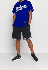 Modrá dres Dodgers s bílým písmem, doplněný černými šortkami s bílým lemováním a logem Nike. Černé sportovní boty se zelenými akcenty.