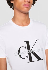 Mladá osoba nosiace biele tričko s veľkým čiernym "CK" a sivým "Calvin Klein Jeans" textom na prednej strane.