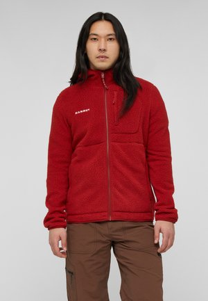 Mann mit langen schwarzen Haaren, der eine rote Fleecejacke mit Frontreißverschluss und braune Cargohosen trägt, steht vor einem schlichten grauen Hintergrund.