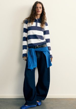 Femme portant une chemise à manches longues rayée bleu et blanc, un jean large foncé, des ballerines bleues et un pull rayé bleu noué autour de la taille.