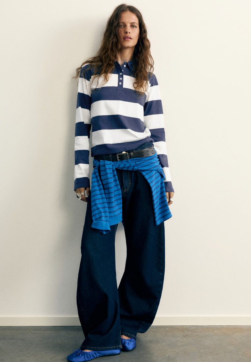 Femme portant une chemise à manches longues rayée bleu et blanc, un jean large foncé, des ballerines bleues et un pull rayé bleu noué autour de la taille.
