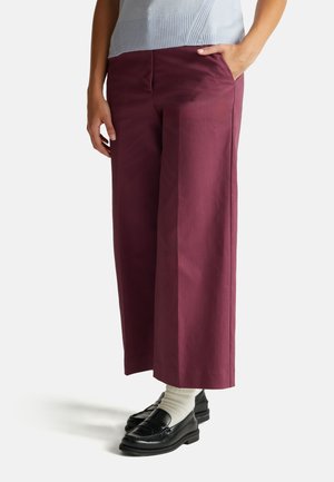 Bordeauxfarbene weit geschnittene Hose aus Baumwolle, mit seitlichen Taschen und glatter Textur, kombiniert mit schwarzen Slippern und weißen Socken.