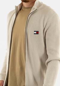 Homme portant un pull zippé côtelé beige par-dessus un t-shirt marron clair, avec le logo Tommy Jeans sur la poitrine à gauche.