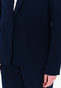 Giacca su misura blu navy con texture liscia. Presenta una tasca singola sul lato sinistro e rever strutturati.