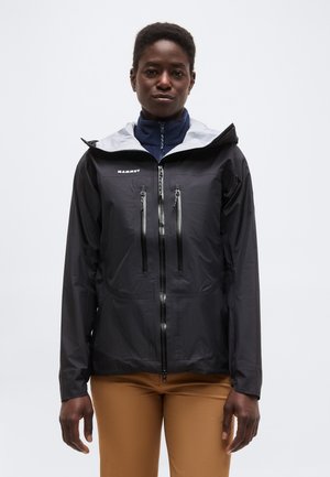 Dospelá osoba v čiernej nepremokavej bunde s vertikálnymi zipsami na hrudi a bielou kapucňou, v kombinácii so svetlohnedými nohavicami, stojaca na bielom pozadí.