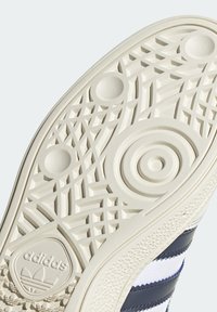 Podešev tenisky Adidas z texturované gumové hmoty s geometrickými vzory a kulatým logem na spodní straně. Převládající krémová barva.