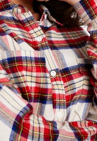 Camicia di flanella a quadri rossa, blu e gialla con bottoni bianchi e colletto a punta. Presenta una texture morbida e motivi orizzontali e verticali ben visibili.