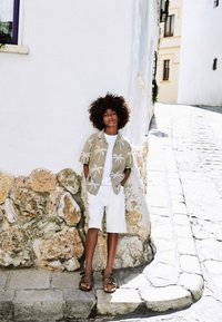 Camicia verde chiaro a maniche corte con motivo di palme bianche, t-shirt bianca, pantaloncini bianchi e sandali marroni, in piedi contro un muro di pietra.