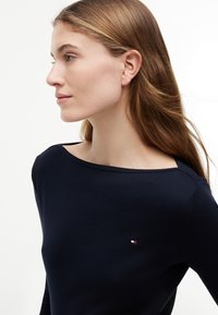 Junge Frau mit langen hellbraunen Haaren, die ein eng anliegendes marineblaues Oberteil mit einem kleinen rot-weiß-blauen Logo auf der Brust trägt.