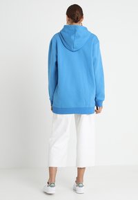 Blå hoodie av mjukt tyg, med huva och ribbade ärmslut, i kombination med vita kortbyxor och sneakers med gröna accenter.