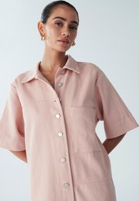Jasnoróżowa sukienka koszulowa z krótkim rękawem, kołnierzykiem, kieszenią z przodu i srebrnymi zatrzaskami. Wykonana z teksturowanej tkaniny o luźnym kroju.
