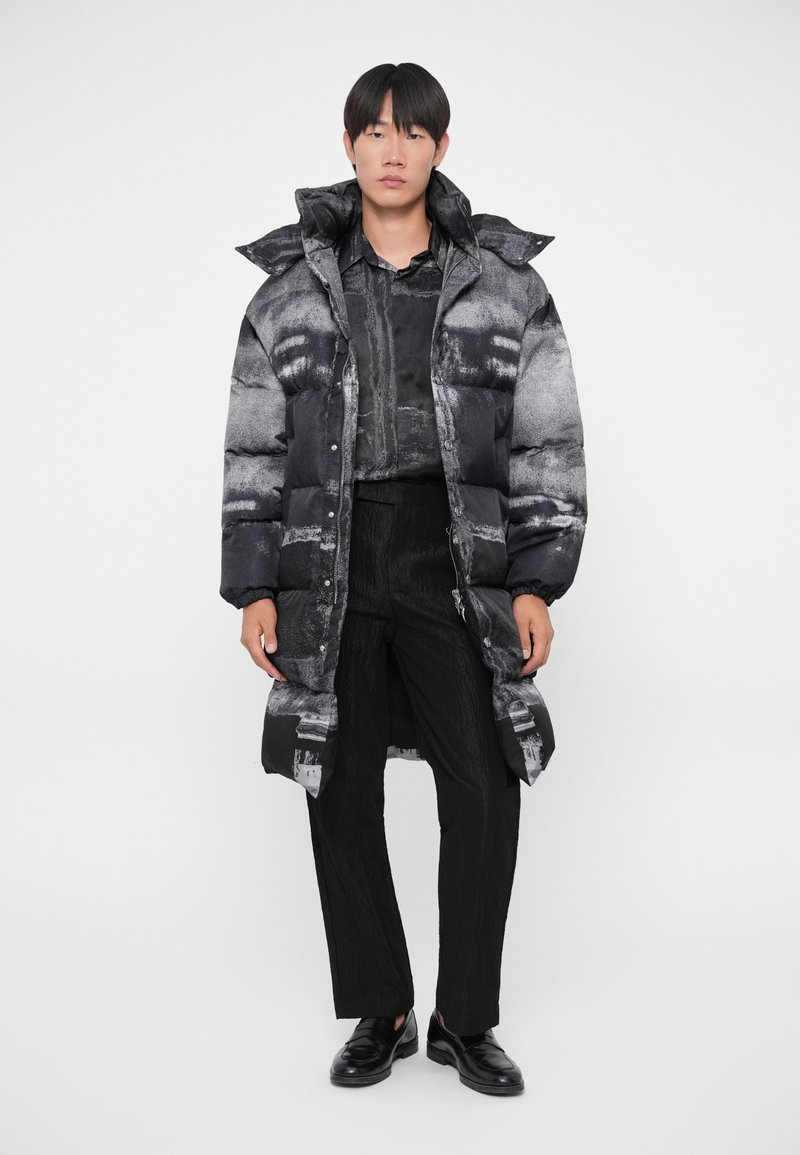 Puffjakke med et sort og gråt abstrakt mønster, der har høj krave, trykknapper og elastiske manchetter, båret over sorte teksturerede bukser.