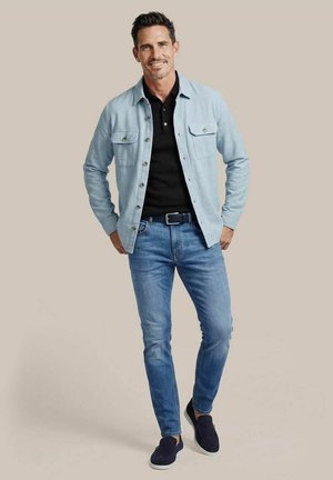 Man draagt lichtblauw overshirt, zwarte polo, blauwe spijkerbroek, zwarte riem en navy slip-on schoenen, staat met handen in de zakken op een beige achtergrond.