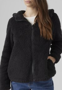 Kvinna som bär en svart zip-up fleecejacka med huva och en hand i fickan, över en vit ribbad topp och blå jeans.