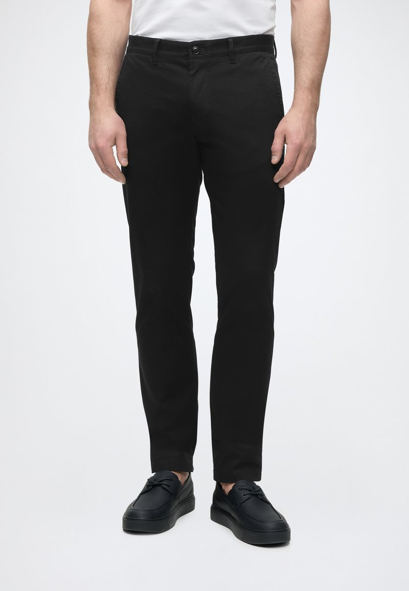Mann trägt schmale schwarze Hose und schwarze Schnürschuhe, steht vor einem einfarbigen hellen Hintergrund.