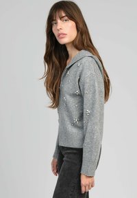 Haut à capuche gris en tricot avec un ourlet côtelé, présentant des embellissements décoratifs et une coupe décontractée. Le tissu texturé met en valeur les détails.