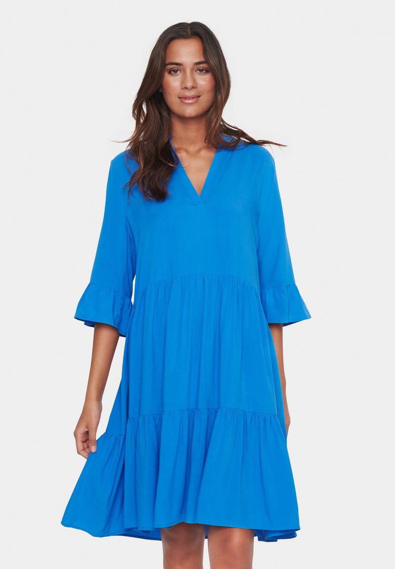 Rochie albastră, până la genunchi, dintr-un material moale, cu decolteu în V, mâneci 3/4 și manșete cu volane. Prezintă un design flared cu straturi la tiv.