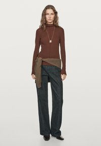 Brauner, figurbetonter Langarm-Turtleneck, kombiniert mit weit geschnittenen, dunklen Jeans. Ein brauner Pullover, der um die Taille gebunden ist, sorgt für einen Kontrast. Detail mit silberner Halskette.