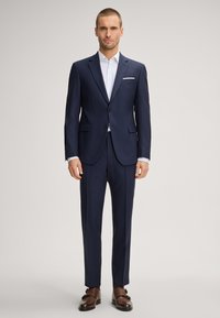 Abito blu navy con vestibilità sartoriale, caratterizzato da una giacca a due bottoni, pantaloni con piega piatta e scarpe di pelle marrone. Camicia bianca texturizzata sotto.