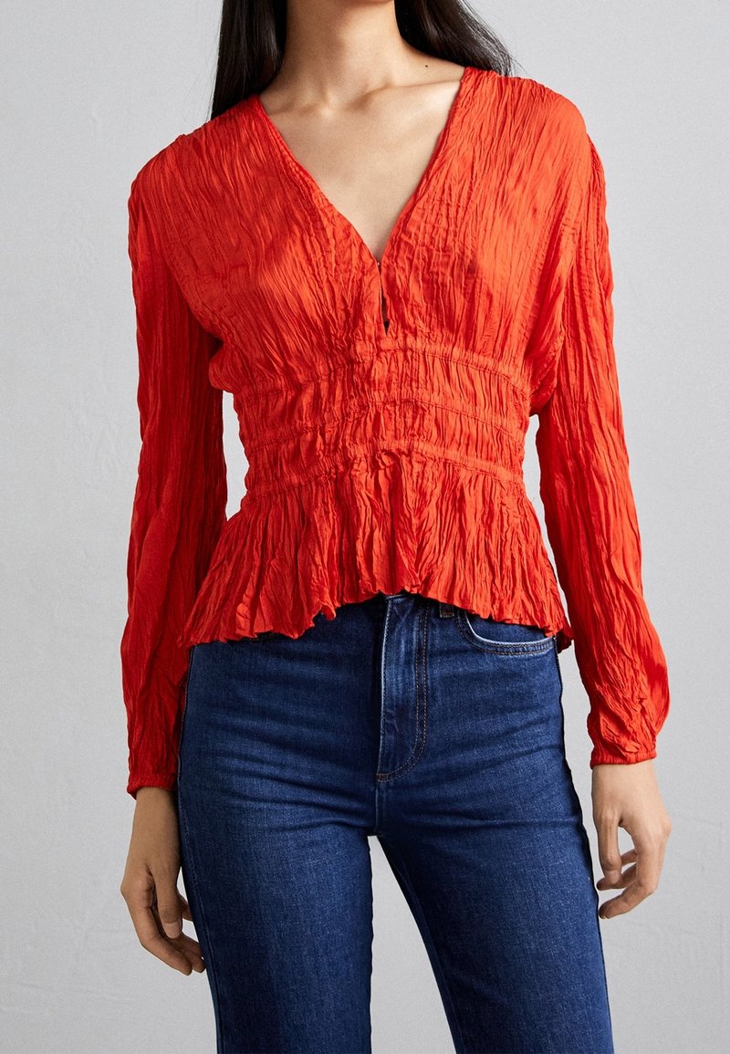Femme portant un chemisier rouge à manches longues, froissé, avec un col en V et un jean bleu foncé taille haute, sur fond clair uni.