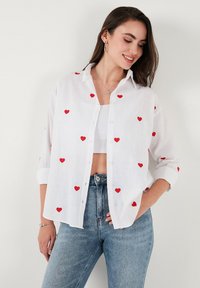 White button-up shirt made of lightweight fabric, featuring red heart embroidery scattered across the surface. Rolled sleeves and relaxed fit.