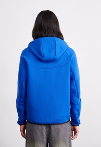Sweatshirt azul com capuz estruturado, tecido texturizado e barra preta em contraste. Apresenta costuras detalhadas nas costas e mangas longas.