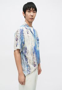 Koszula z krótkim rękawem w kremowym kolorze z niebiesko-brązowymi abstrakcyjnymi wzorami, posiada kołnierzyk i kieszonkę na piersi, wykonana z gładkiej tkaniny.