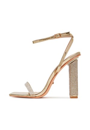 Złota sandałka na szpilce z paskiem zdobionym kryształami i obcasem typu słupek, z metalicznym wykończeniem i kwadratowym noskiem.